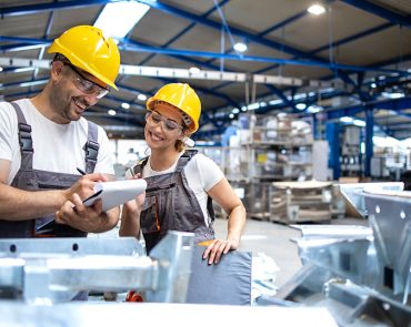 Two workers in a manufacturer’s factory, wearing yellow hard hats and safety glasses, smile as they review documents on a clipboard. Surrounded by industrial equipment and machinery inside a spacious, well-lit warehouse, the setting reflects a clear view of modern efficiency.