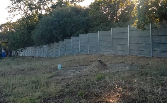 A long, gray ClamberPrufe Clear View Fencing panel runs parallel to a patch of dry, grassy ground with a green bucket and a shovel resting on a dirt mound. Dense trees and shrubs grow behind the fence.