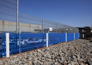 A secure parking area is enclosed by blue metal fencing and tall clear view fencing topped with barbed wire, beside a rocky ground and under a clear blue sky.