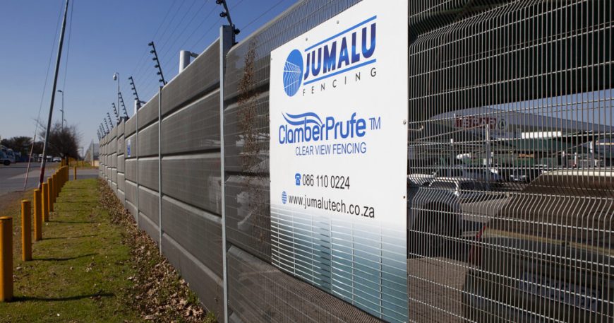 A tall metal fence with a sign that reads Jumalu Fencing, Chamber Prufe Clear View Fencing. This clear view security fencing stands by a road with yellow posts, reflecting nearby buildings. Contact details are listed for inquiries.