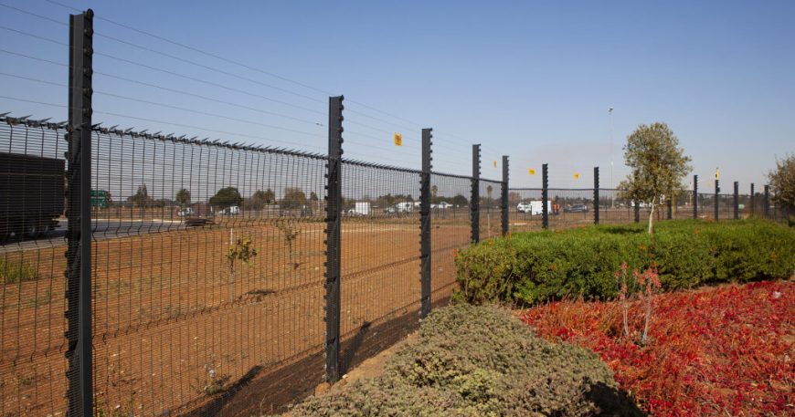 A clear view fence stretches alongside a road with a barren field on one side and a landscaped area featuring green and red foliage offering a clear view on the other. Trees and vehicles can be seen in the background under the pristine blue sky.