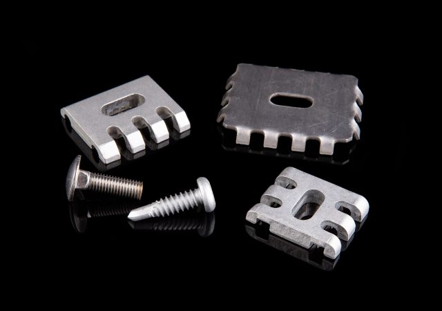 Four metal mechanical components, including two threaded screws and two rectangular blocks with grooves, are displayed on a reflective black surface, reminiscent of the precision found in Clearview fence designs.