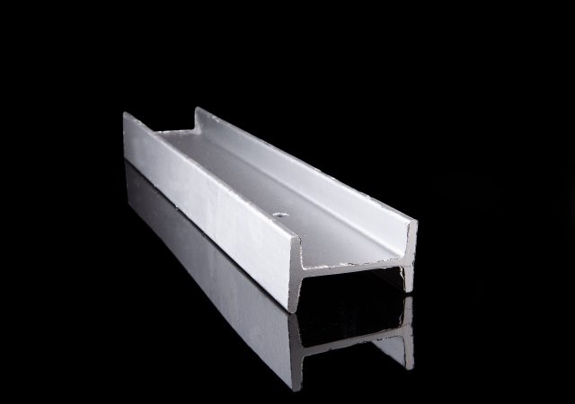 A single aluminum C-channel beam, resembling the structure of Clearview fencing, is displayed on a reflective black surface. The beam is positioned diagonally with one end closer to the viewer, showcasing its U-shaped cross-section.