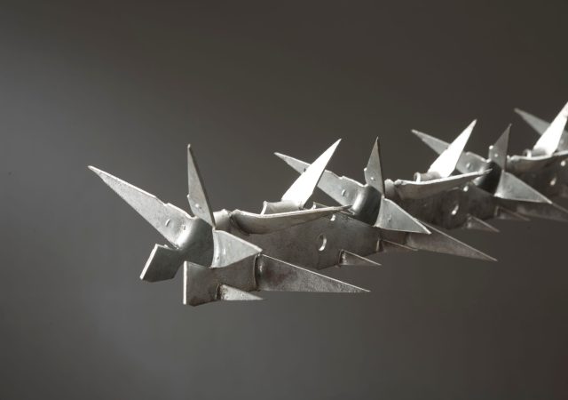 Close-up of razor wire against a dark, blurred background, the angular metallic spikes of the clambrufe clearview fencing reflecting light. This creates a stark and intense image.